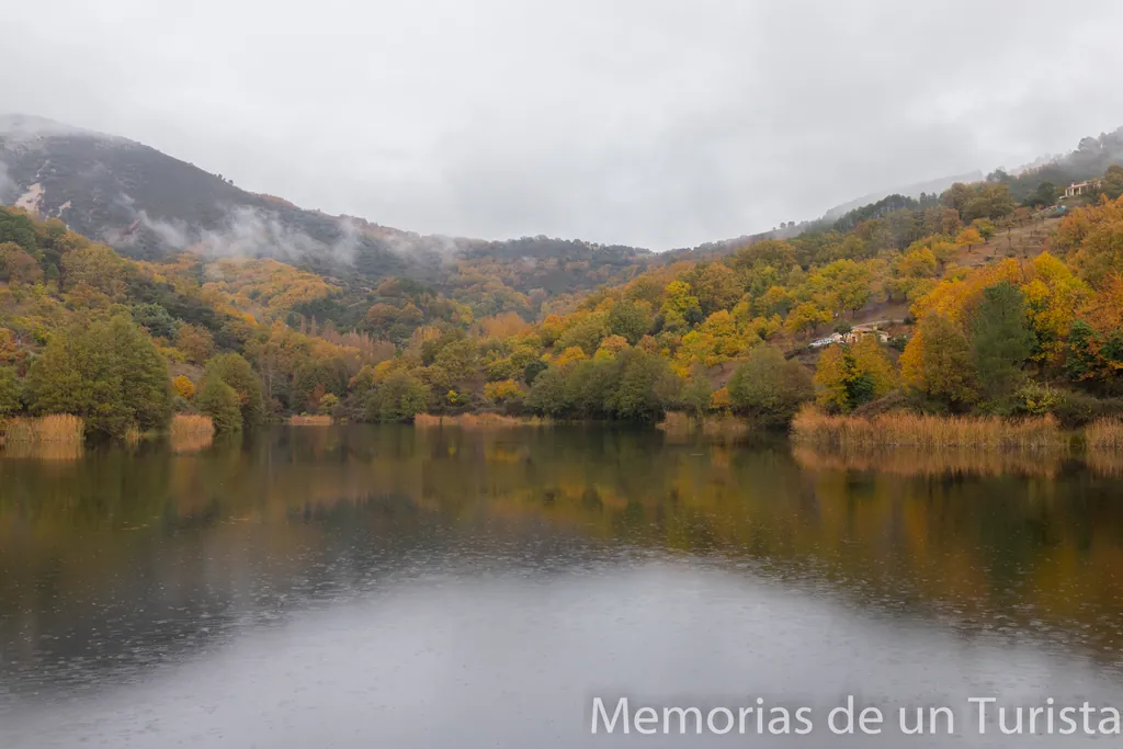Paisaje otoñal de la presa en la Ruta de los Molinos, Guadalupe. El agua tranquila refleja un bosque de castaños y robles con hojas amarillas y ocres, bajo un cielo nublado con jirones de niebla descansando sobre las montañas