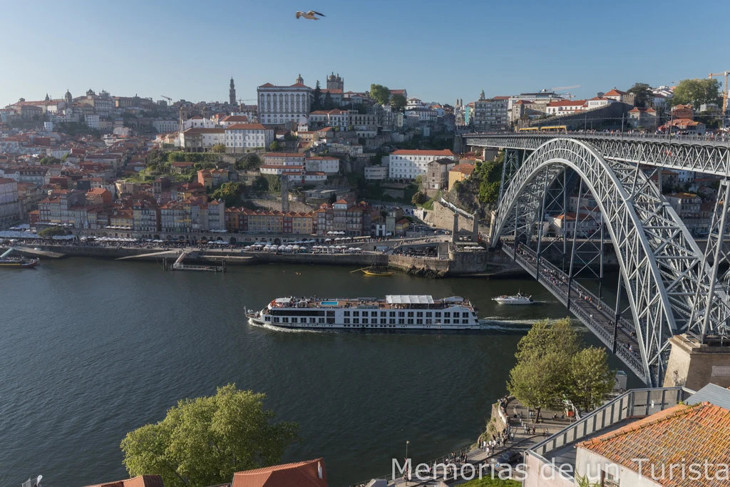 Vista panorámica del río Duero en Oporto con el puente de Don Luis I a la derecha, un crucero navegando y las coloridas casas de la Ribeira al fondo bajo un cielo despejado