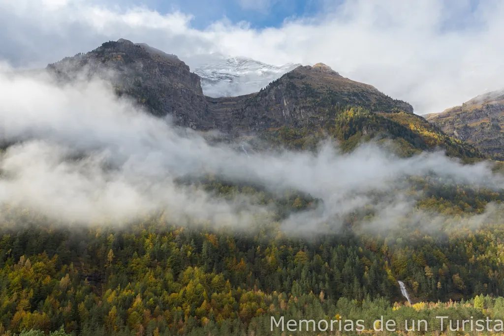 Vista de las montañas del Valle de Pineta desde la carretera hacia el Parador de Bielsa, mostrando laderas boscosas con colores otoñales, jirones de niebla blanca y cumbres escarpadas parcialmente nevadas bajo un cielo nublado