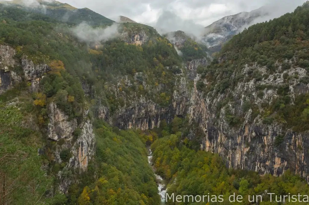 Vistas panorámicas desde los Miradores de Revilla, Huesca, mostrando un profundo cañón de paredes rocosas escarpadas, bosques de pinos y vegetación de fondo de valle con un río, bajo un cielo nublado y con jirones de niebla