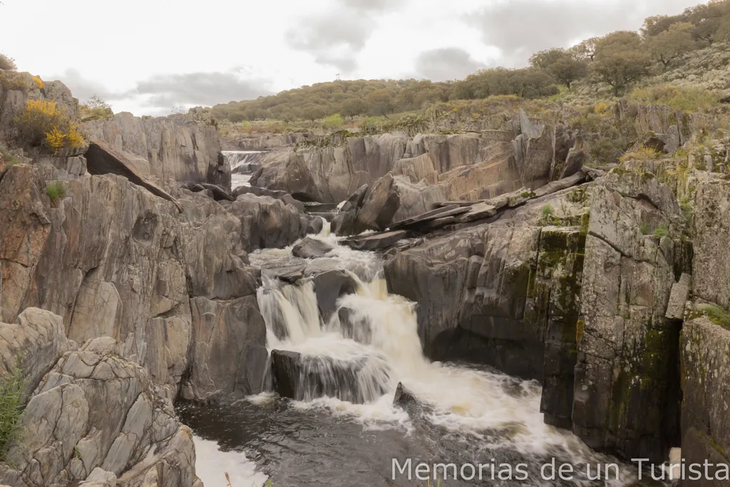 Vista de una cascada escalonada en el río Almonte, Jaraicejo. El agua fluye con fuerza entre grandes formaciones de rocas verticales y afiladas bajo un cielo nublado, rodeada de la vegetación típica de la dehesa extremeña