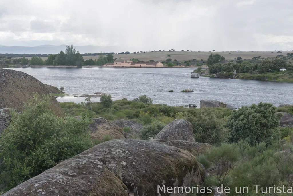 Vista panorámica de la Charca de los Barruecos de Abajo en Malpartida de Cáceres, Extremadura. En primer plano, grandes rocas de granito cubiertas de musgo y vegetación baja. Al fondo, tras el agua, se distingue el edificio histórico de paredes claras que alberga el Museo Vostell Malpartida, bajo un cielo nublado que proyecta reflejos en la superficie de la charca